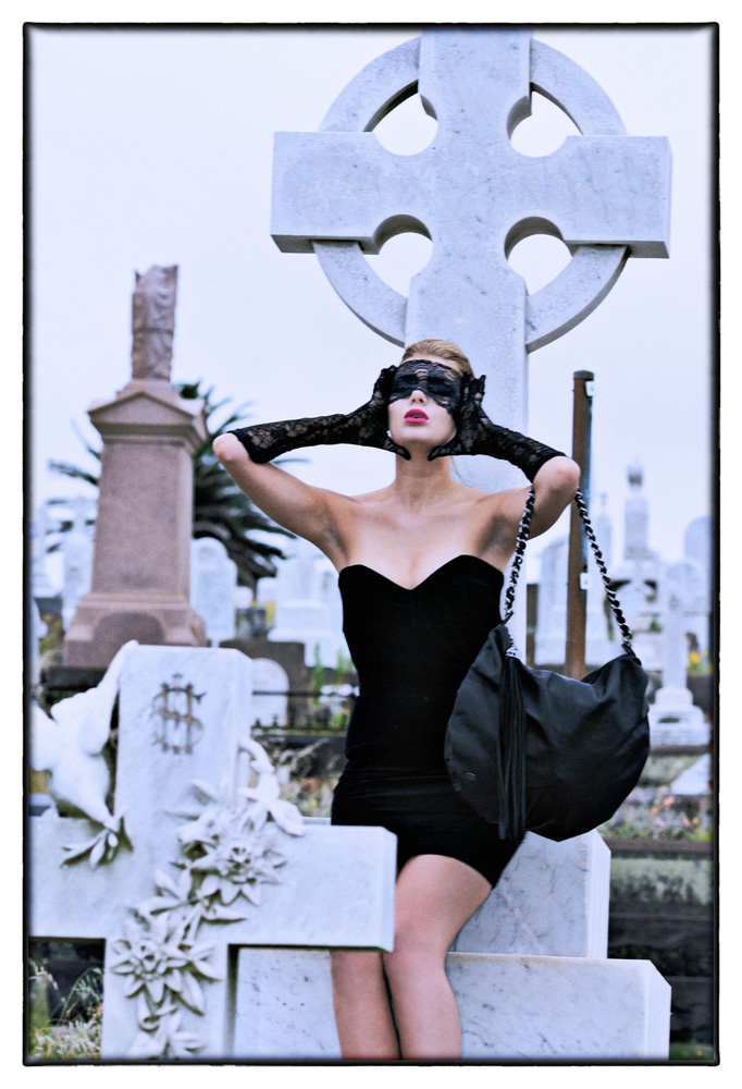 A woman in mourning dress and lace mask cries out in front of a marble cross in a cemetery.