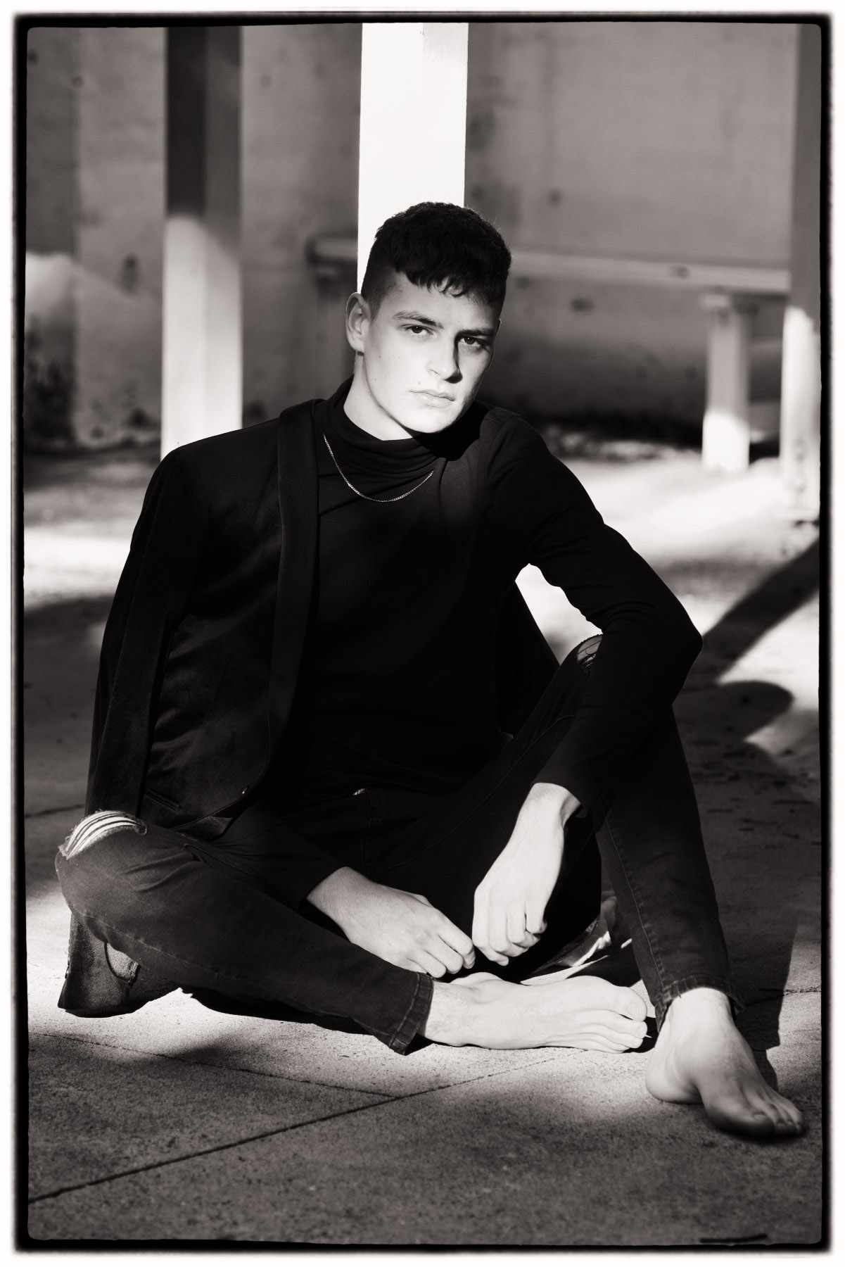 Male model seated on ground in strong light, black and white portfolio photoshoot on location.