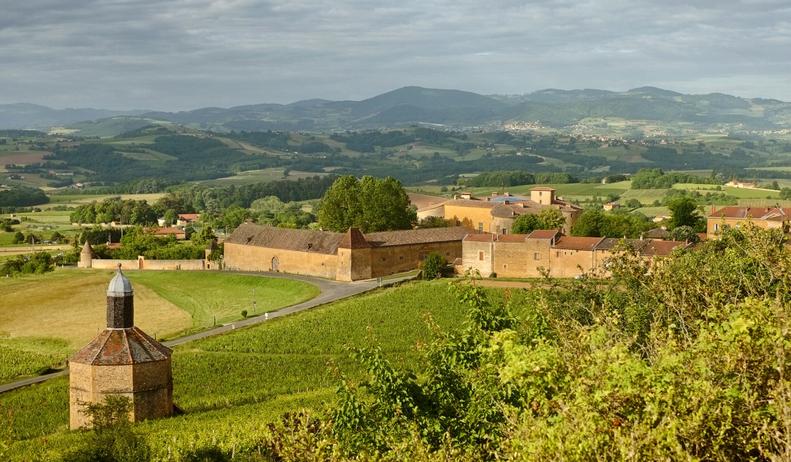 A scenic landscape of hills, vineyards, the Pigeonnier and Château de Bagnols. Part of the Beaujolais wine region, Arrondissement: Villefranche-sur-Saône, France. Travel photography by Kent Johnson.