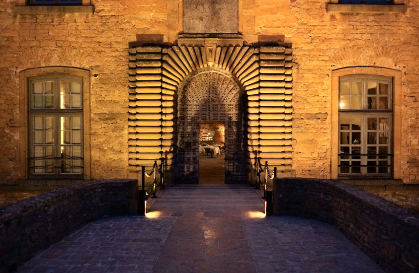 The drawbridge, stonework and studded door of Château de Bagnols at night. Travel photography by Kent Johnson.
