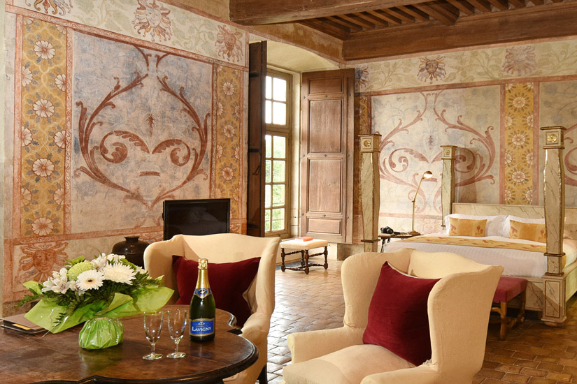 Château hotel suite; the expansive main room decorated with mural and stenciled walls, furnished with dining and occasional tables, wing backed chairs and four poster bed. Luxury hotel interior photography by Kent Johnson.