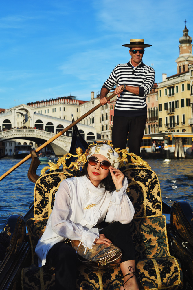 Vivienne riding in a Gondola wearing a white pirate blouse, being rowed down the Grand Canal, Venice in last light. Fashion Photography by Kent Johnson