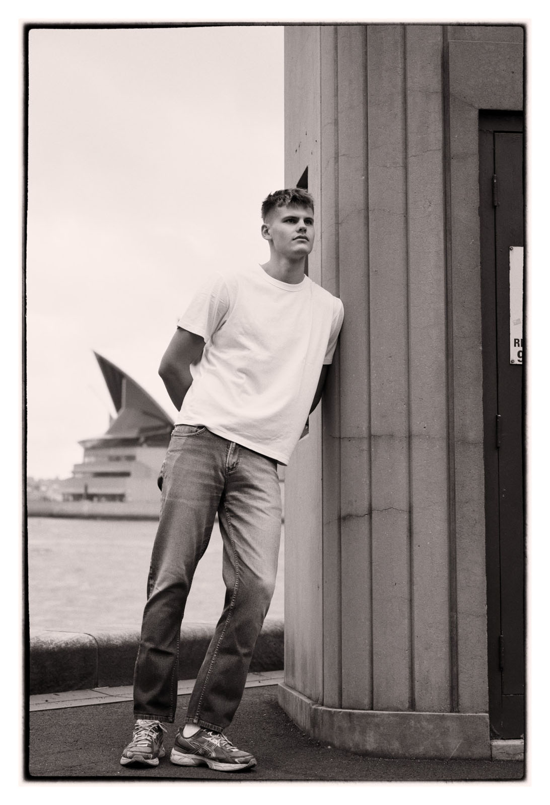 A fashion portrait and travel momory image of a young male traveller, Black and white photograph with the Sydney Opera House as a backdrop. Portrait by Kent Johnson Photography, Sydney, Australia.