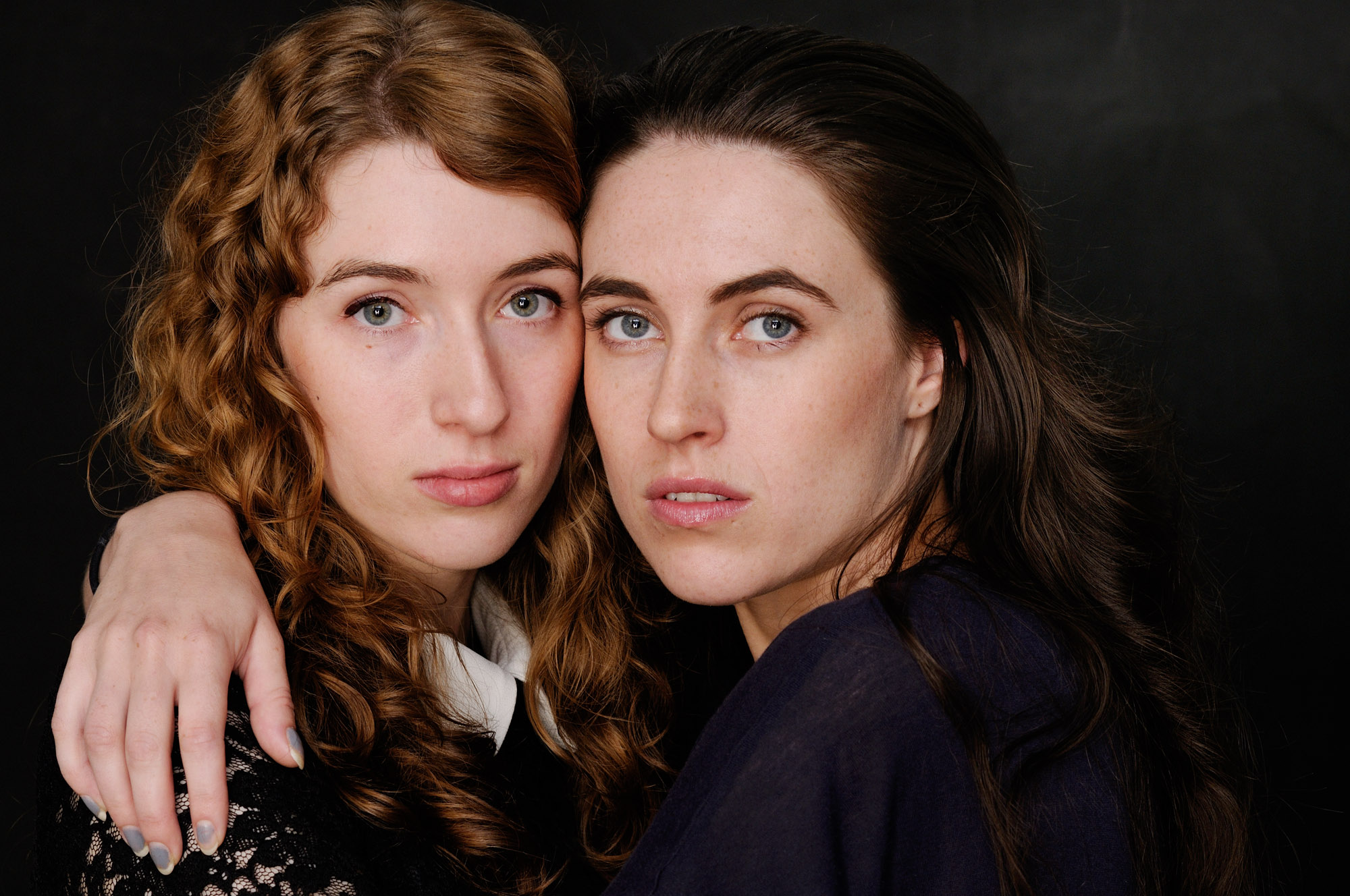 Two sisters embrace in a tight studio portrait photographed against a dark background. Photographed by Kent Johnson.